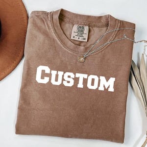 a brown t-shirt with the word "CUSTOM" printed on it, along with a brown hat and a plant.