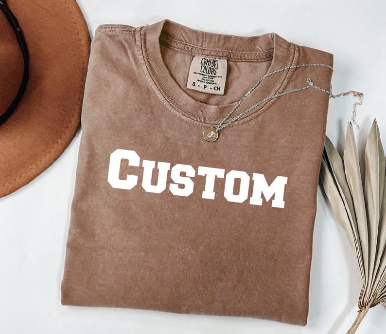 a brown t-shirt with the word "CUSTOM" printed on it, along with a brown hat and a plant.