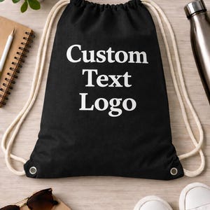 May include: A black drawstring backpack featuring the white text Custom Text Logo printed in the centre. The bag has thick off-white rope straps that cinch the top closed and silver metal grommets at the bottom corners for the cord attachments.