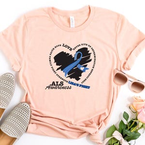 May include: A peach-colored t-shirt featuring a black heart graphic with a blue awareness ribbon. The design includes the text LOVE, FAITH, HOPE, NEVER GIVE UP, DETERMINATION, FAMILY, FIGHT, STRENGTH, CURE, COURAGE, ALS AWARENESS, and LISA'S FIGHT.
