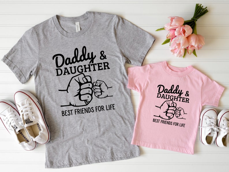 May include: Two gray and pink t-shirts with a black graphic of two fists bumping. The text on the shirts reads "Daddy & Daughter Best Friends For Life".