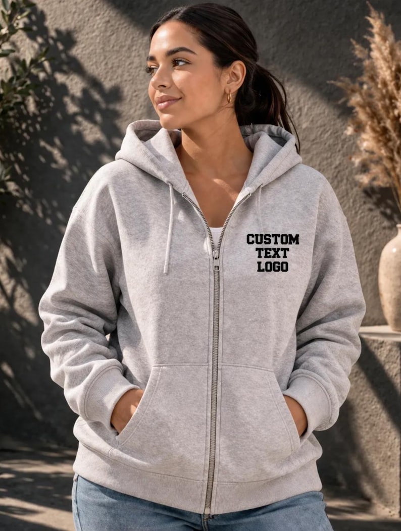 A woman wearing a grey hooded sweatshirt with the text "CUSTOM TEXT LOGO" printed on it stands in front of a gray wall.