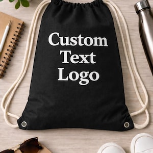 May include: Black drawstring backpack with the words "Custom Text Logo" in white. The bag has cream-colored drawstrings and metal eyelets. Other items in the image include sunglasses, a notebook, a pencil, and white sneakers.