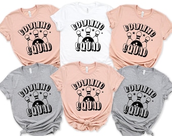 May include: A collection of six short-sleeved t-shirts in white, light pink, and heather gray. Each shirt features a black graphic print of three bowling pins and a bowling ball with the text BOWLING SQUAD arched above and below the graphic.