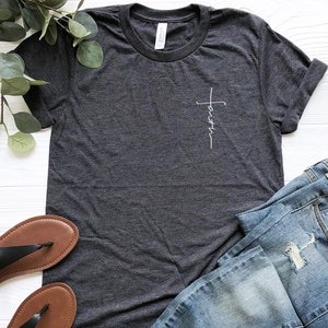 May include: A dark gray t-shirt with a white cross and the word "faith" written vertically on the left side of the chest.
