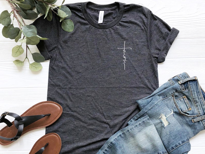 May include: A dark gray t-shirt with a white cross and the word "faith" written vertically on the left side of the chest.