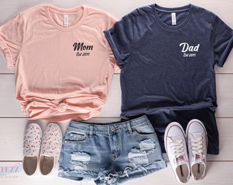 May include: Two t-shirts, one pink and one navy blue, with the words "Mom Est 2011" and "Dad Est 2011" printed on them. The shirts are laid out on a white wooden surface with a pair of white sneakers and a pair of denim shorts.