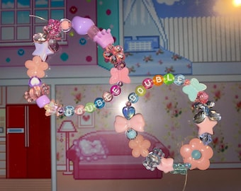 Könnte beinhalten: Ein Schnullerclip mit Perlen, auf dem das Wort "DOUBLE" in bunten Buchstabenperlen geschrieben steht. Der Clip hat rosa, lila und transparente Perlen sowie Blumen-, Stern- und Schmetterlingsanhänger. Der Clip ist für Babys konzipiert.