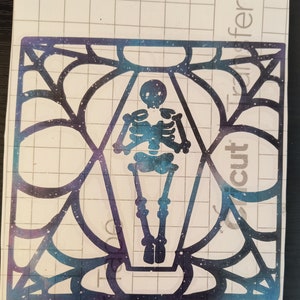May include: A die-cut sticker featuring a skeleton inside a coffin-shaped frame. The skeleton and coffin have a galaxy-themed design with blue, purple, and white colors. The frame has a spiderweb-like pattern.