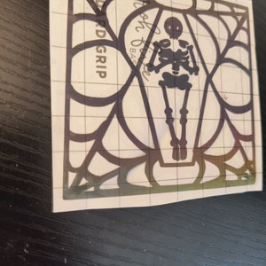 May include: A black skeleton figure inside a black spiderweb design. The design is on a white background with a grid pattern.