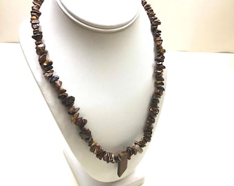 May include: A necklace composed of numerous irregular, polished brown and golden-brown tiger's eye stone chips strung together, featuring a single, larger, rectangular-shaped tiger's eye stone pendant hanging at the center of the strand.