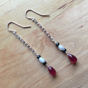 May include: Dangle earrings with silver chain links. Each earring has a white pearl, black beads, and a faceted, teardrop-shaped red gemstone. The earrings are presented on a wooden surface.