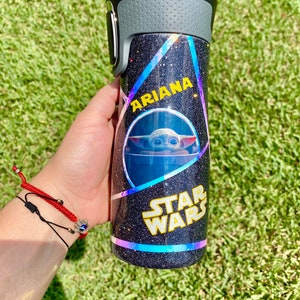 May include: A black and silver tumbler with a glittery finish. The tumbler has a holographic design with a Star Wars logo and a picture of Baby Yoda. The name "Ariana" is written on the tumbler.