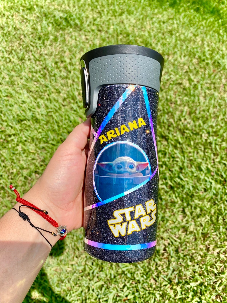 May include: A black and silver tumbler with a glittery finish. The tumbler has a holographic design with a Star Wars logo and a picture of Baby Yoda. The name "Ariana" is written on the tumbler.