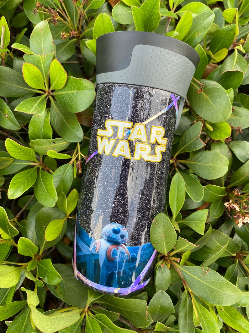 May include: A black and silver travel mug with a glittery finish. The mug features a Star Wars logo and a graphic of the character BB-8.