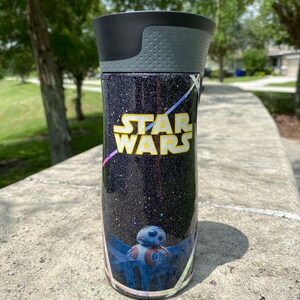 May include: A black and silver tumbler with a glittery black background and a Star Wars design. The design features the text "Star Wars" in yellow and a blue and red droid character.