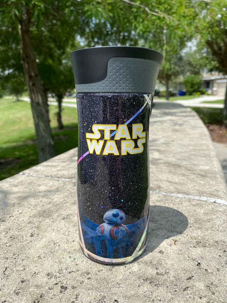 May include: A black and silver tumbler with a glittery black background and a Star Wars design. The design features the text "Star Wars" in yellow and a blue and red droid character.