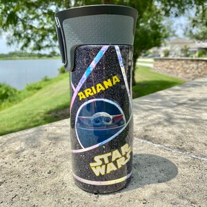May include: A black and silver tumbler with a holographic glitter finish. The tumbler has a black lid with a gray handle. The tumbler has a yellow sticker with the text "Ariana" and a sticker with a character from Star Wars. The tumbler is sitting on a gray surface.