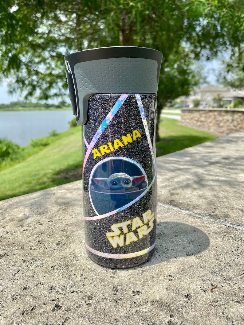 May include: A black and silver tumbler with a holographic glitter finish. The tumbler has a black lid with a gray handle. The tumbler has a yellow sticker with the text "Ariana" and a sticker with a character from Star Wars. The tumbler is sitting on a gray surface.