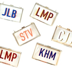 May include: Six clear acrylic clutches with gold hardware and colorful letter monograms. The monograms are "JLB", "LMP", "STV", "CJ", "KHM", and "LMP".