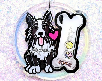 Op de afbeelding: Een zwart-witte border collie met een roze hart en een wit bot met een rode bal, een roze hart en een touwspeelgoed erin. De hond en het bot bevinden zich op een doorzichtige ronde sleutelhanger met een regenboogkleurige, iriserende achtergrond.