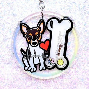 May include: A round, clear acrylic keychain featuring a white dog with brown markings holding a red heart and a bone. The bone has the text "whatta charmer" on it. The keychain has a silver metal ring.