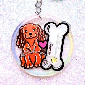 Puede incluir: Un llavero acrílico transparente con un perro de dibujos animados Cavalier King Charles Spaniel con un corazón rosa, un hueso blanco y el texto "whatttacharmer". El llavero tiene un cierre y una cadena plateados.