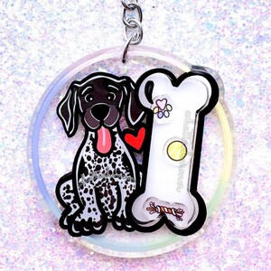 May include: A clear keychain with a black and white cartoon German Shorthaired Pointer dog and a white bone with red hearts and a tennis ball. The keychain is attached to a silver key ring.