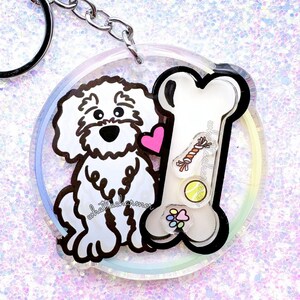 以下が含まれることがあります： ピンクのハート、骨、犬のおもちゃが付いた白い漫画の犬が特徴の透明なアクリルキーホルダー。キーホルダーにはシルバーの留め金とリングが付いています。「whatthecharmer」というテキストが表示されます。
