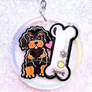 May include: A clear acrylic keychain featuring a black and tan dog with a pink heart, a white bone, and the text "shellacharmers". The keychain has a silver keyring and clasp. The bone has a paw print, a rainbow, a yellow ball, and a red and brown rope toy.