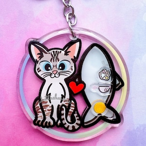 Può includere: Un portachiavi in ​​acrilico trasparente con un gatto dei cartoni animati con pelliccia grigia e bianca e un cuore rosso. Il gatto tiene un pesce bianco con un tuorlo d'uovo giallo all'interno. Il portachiavi ha un anello arcobaleno attorno al bordo.