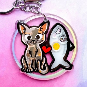 May include: A clear acrylic keychain featuring a cartoon cat and a fish. The cat has a camouflage pattern with a red heart. The fish has a yellow yarn ball and a paw print. The keychain has a silver clasp and ring.