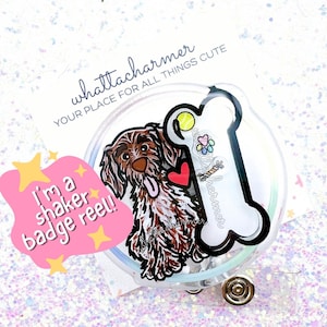 Può includere: Un rotolo per badge in plastica trasparente con un'illustrazione di un cane marrone e bianco, un osso, una palla da tennis e un cuore rosso. Il testo "I'm a shaker badge reel!" è su una nuvola rosa con stelle gialle. Il testo "whattacharmr YOUR PLACE FOR ALL THINGS CUTE" è su uno sfondo bianco.