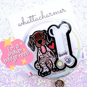 Può includere: Un portabadge trasparente con un'illustrazione di un cane marrone e bianco, un osso e un cuore. Il testo "I'm a shaker badge reel!" è su uno sfondo rosa con stelle gialle. Il testo "whattacharmer" è su uno sfondo bianco.