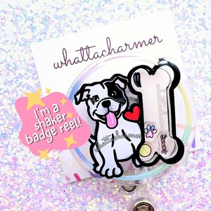 May include: A clear plastic badge reel with a white and black cartoon dog and a bone. The dog has a red heart on its chest and is sticking its tongue out. The text "I'm a shaker badge reel!" is on a yellow star-shaped speech bubble.