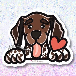 May include: A brown and white German Shorthaired Pointer dog with a red heart in its mouth. The dog is peeking over a white surface with the text "whattacharmer" on it.