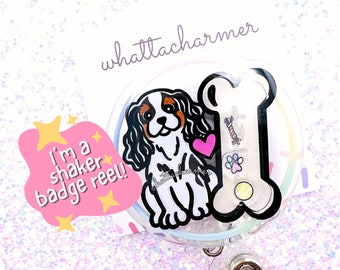 May include: A clear plastic badge reel with a cartoon Cavalier King Charles Spaniel and a bone. The dog is white with brown and black markings. The text "I'm a shaker badge reel!" is on a pink cloud with yellow stars.