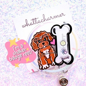 May include: A clear plastic badge reel with a brown and white cartoon dog and a black bone. The text "I'm a shaker badge reel!" is on a pink and yellow sticker. The text "whattacharmer" is on a white sticker.