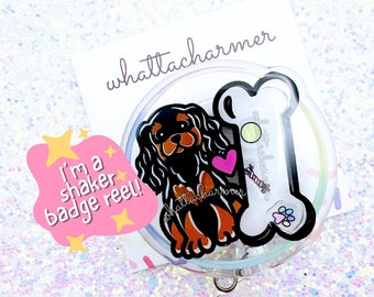 May include: A clear plastic badge reel with a black and brown Cavalier King Charles Spaniel dog illustration, a pink heart, and a bone with a paw print. The text "I'm a shaker badge reel!" is on a pink sticker with yellow stars.