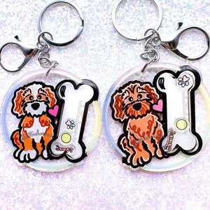 May include: Two round, iridescent keychains featuring a white bone and a cartoon doodle of a brown and white dog. The dog is holding a pink heart. The bone has a tennis ball, a rope toy, and paw prints on it.