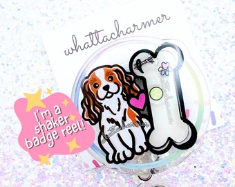 May include: A clear shaker badge reel featuring a Cavalier King Charles Spaniel illustration with a pink heart. The reel also includes a bone-shaped charm and the text "I'm a shaker badge reel!!". The background is a glittery white.