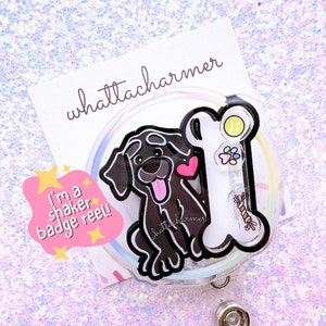 Peut inclure: Un badge rétractable rond transparent avec un labrador noir, un cœur rouge et un os en jouet. Le badge rétractable a un motif arc-en-ciel et le texte "I'm a shaker badge reel!" est sur une étoile rose. Le texte "whattacharmer" est sur une carte blanche.