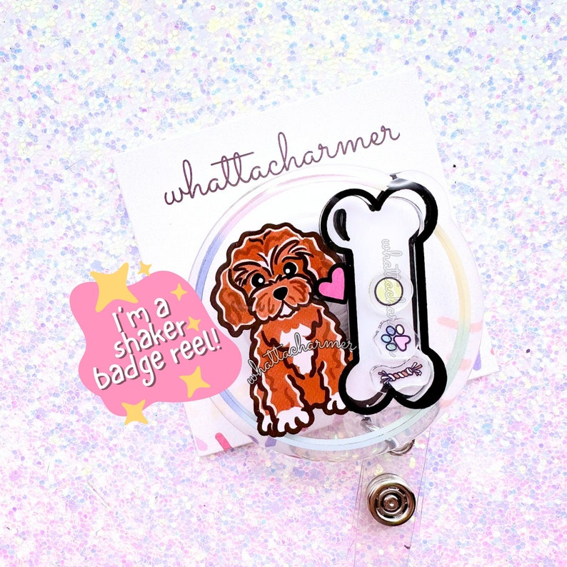May include: A clear plastic badge reel featuring a brown and white cartoon dog and a black bone. The text "I'm a shaker badge reel!" is on a pink and yellow sticker. The text "whattacharmer" is on a white sticker.