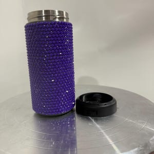 May include: A purple stainless steel tumbler with a sparkly, glittery finish. The tumbler is sitting on a silver surface.