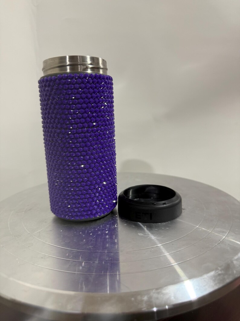 May include: A purple stainless steel tumbler with a sparkly, glittery finish. The tumbler is sitting on a silver surface.