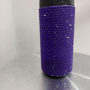 May include: A purple Yeti tumbler covered in rhinestones. The tumbler has a black lid with the Yeti logo.