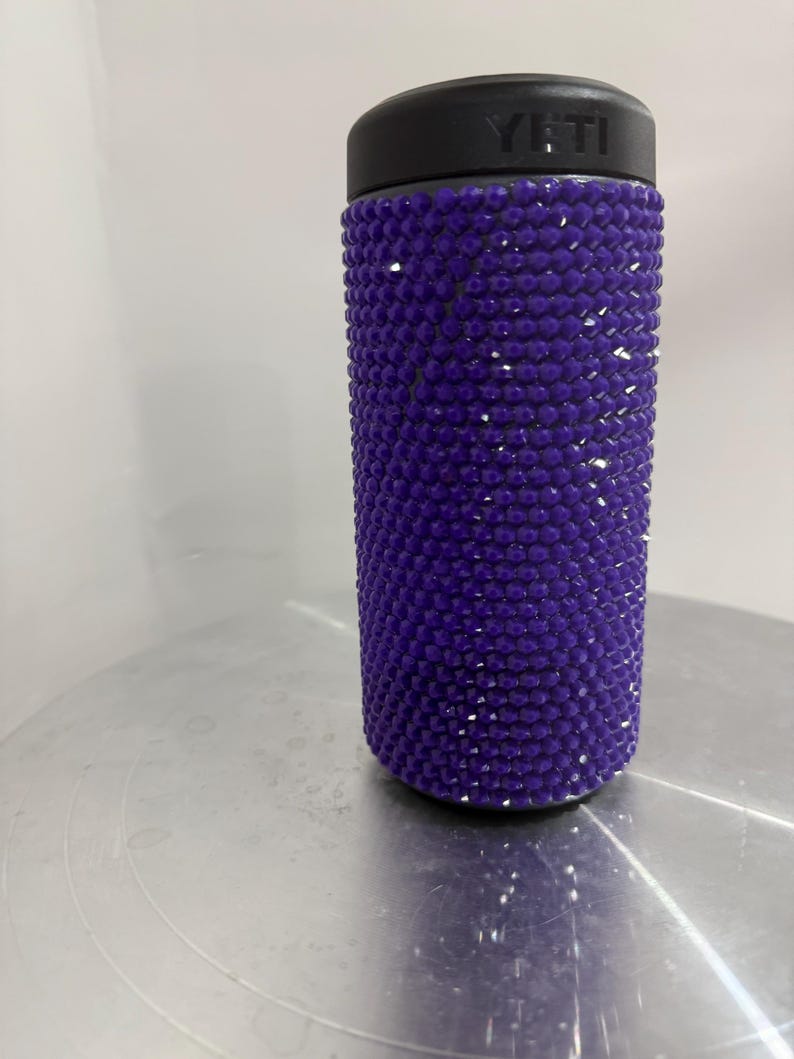 May include: A purple Yeti tumbler covered in rhinestones. The tumbler has a black lid with the Yeti logo.