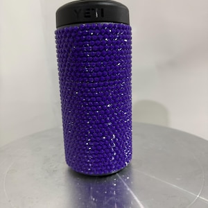 May include: Purple Yeti tumbler covered in rhinestones. The tumbler has a black lid with the word "YETI" printed on it.