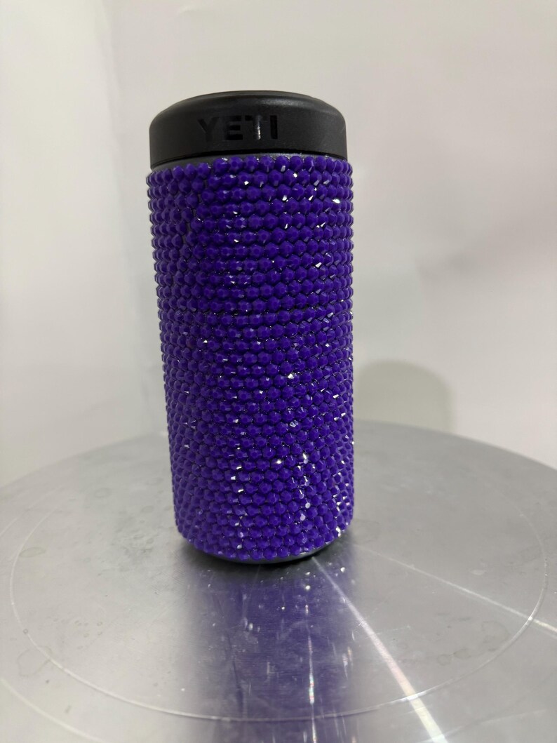 May include: Purple Yeti tumbler covered in rhinestones. The tumbler has a black lid with the word "YETI" printed on it.