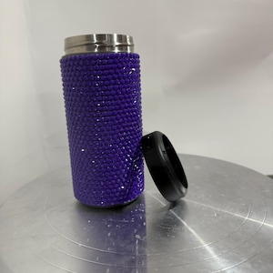 May include: A purple travel mug with a stainless steel lid and a sparkly purple finish. The mug is sitting on a silver surface.